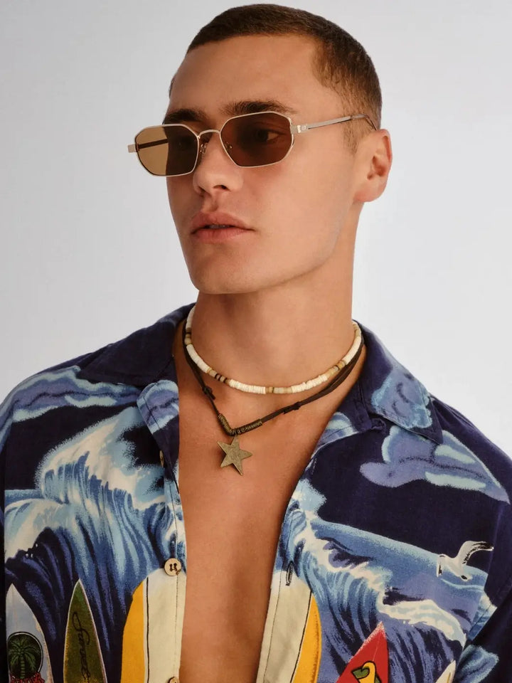 A studio portrait of a man looking to the side while wearing the geometric Le Specs Mythic - Gold Light Brown Mono sunglasses and a blue wave-patterned shirt | Twentyseven Toronto