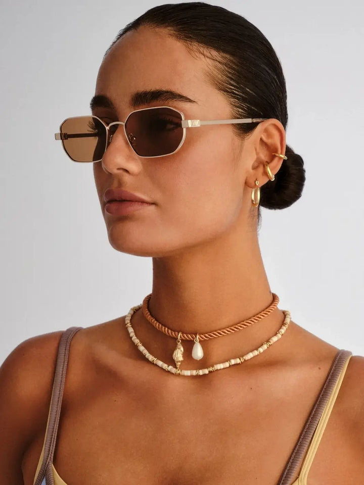 A three-quarter studio portrait of a woman looking to the left to showcase the slim metal temple arm design of the Le Specs Mythic - Gold Light Brown Mono sunglasses | Twentyseven Toronto