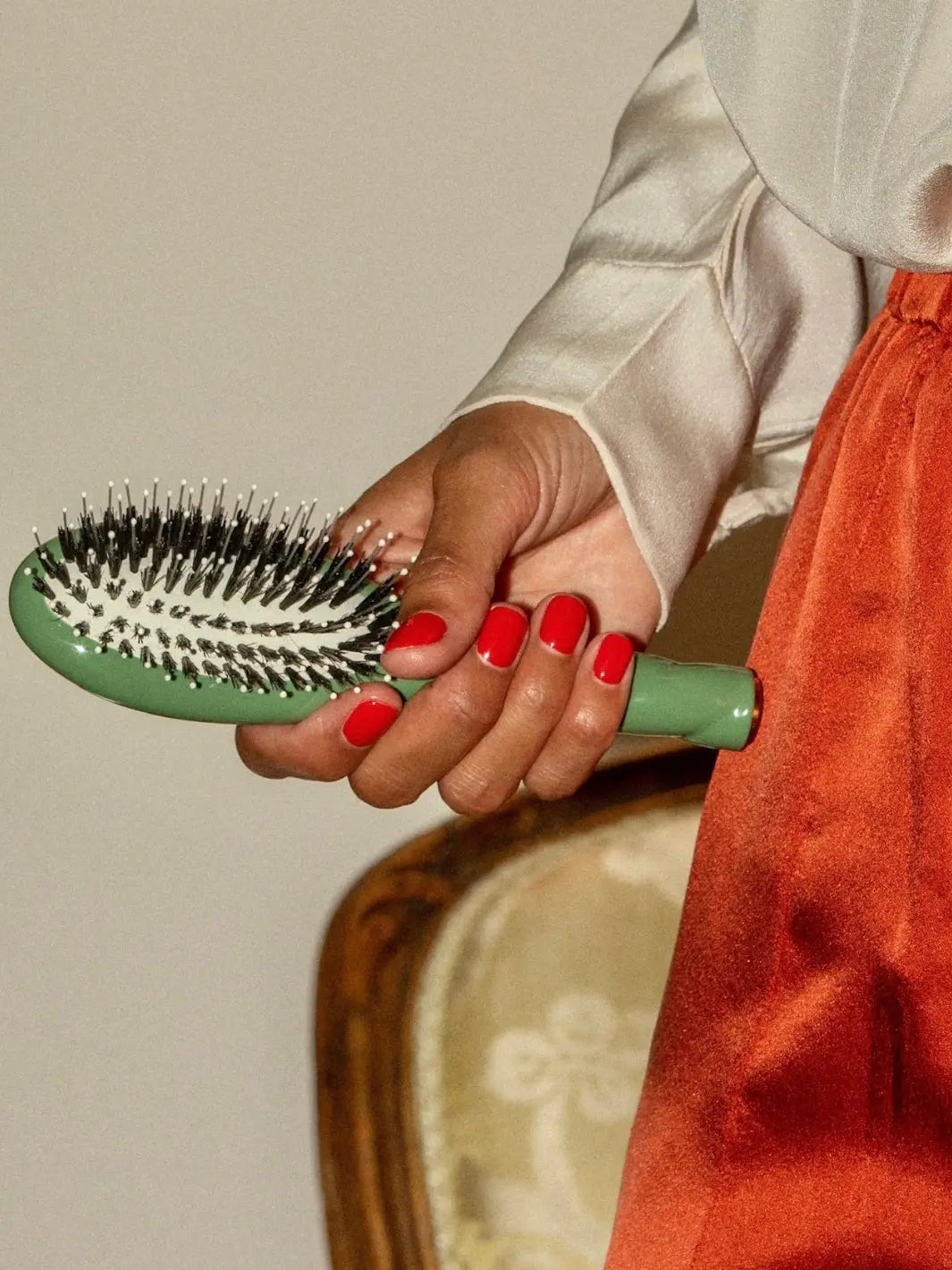 Person with red manicured nails holds the La Bonne Brosse N.03 Small The Essential Softness & Sensitive Scalp Brush in Almond Green while wearing a silk cream blouse and a orange skirt | Twentyseven Toronto