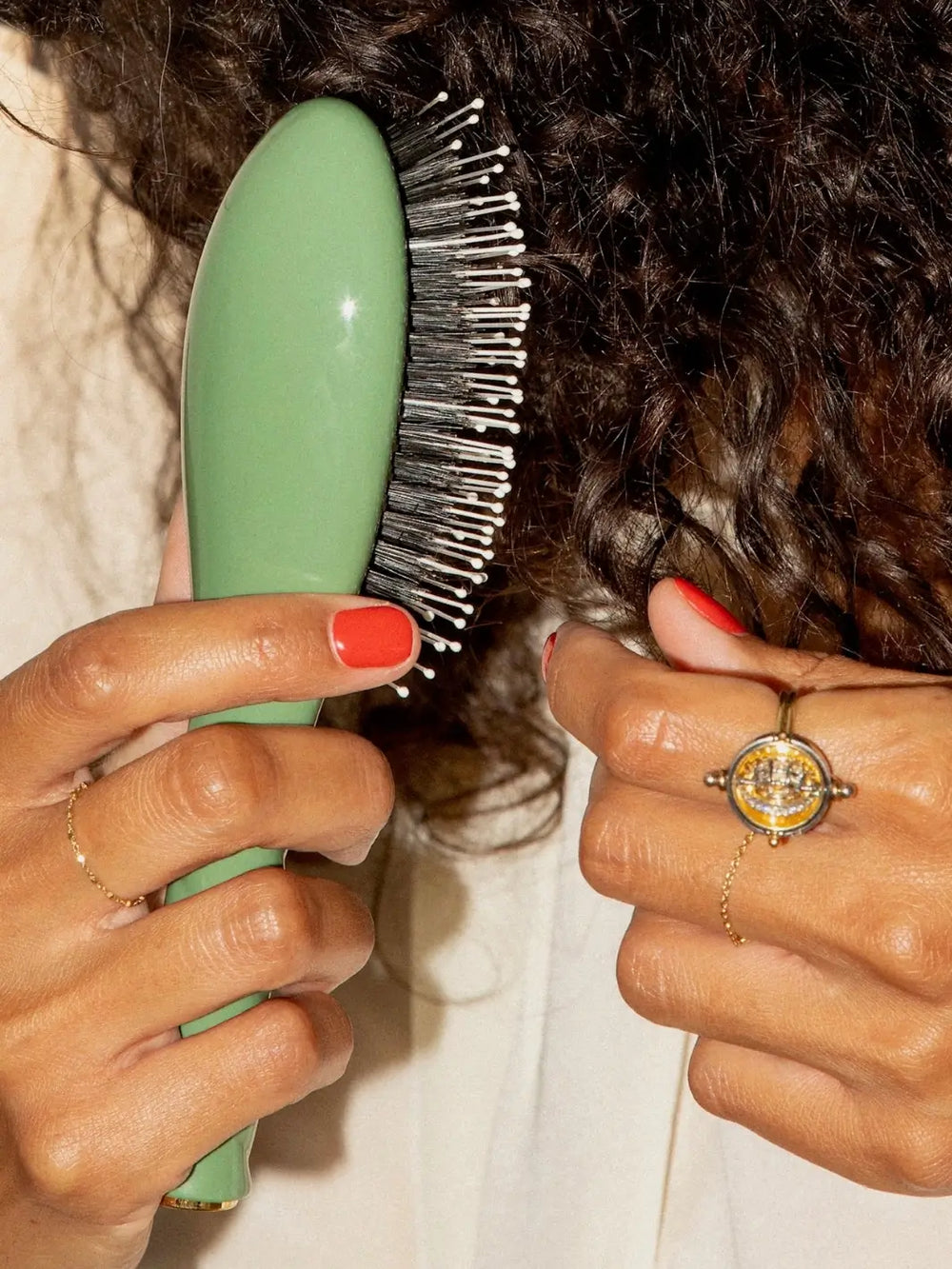 Hands with bright red nails uses the La Bonne Brosse N.03 Small The Essential Softness & Sensitive Scalp Brush in Almond Green to brush through a section of dark, curly hair | Twentyseven Toronto