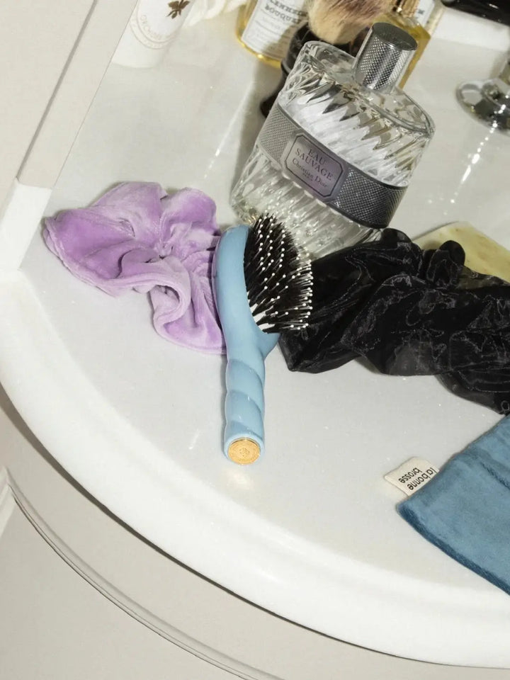 La Bonne Brosse N.03 Small The Essential Softness & Sensitive Scalp Brush in Light Blue sits on a white vanity alongside a bottle of fragrance and purple and black velvet hair scrunchies | Twentyseven Toronto