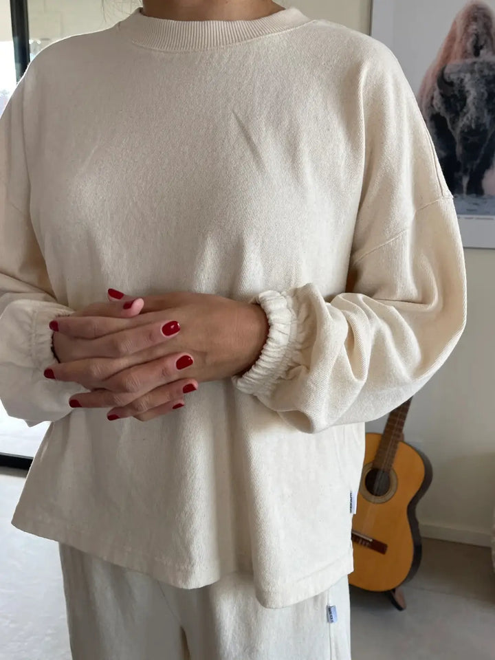 Person wearing a cream 'Naturelle Tee' sweater in 'Naturel' from Le Bon Shoppe with red nail polish, standing indoors with a guitar in the background | Twentyseven Toronto