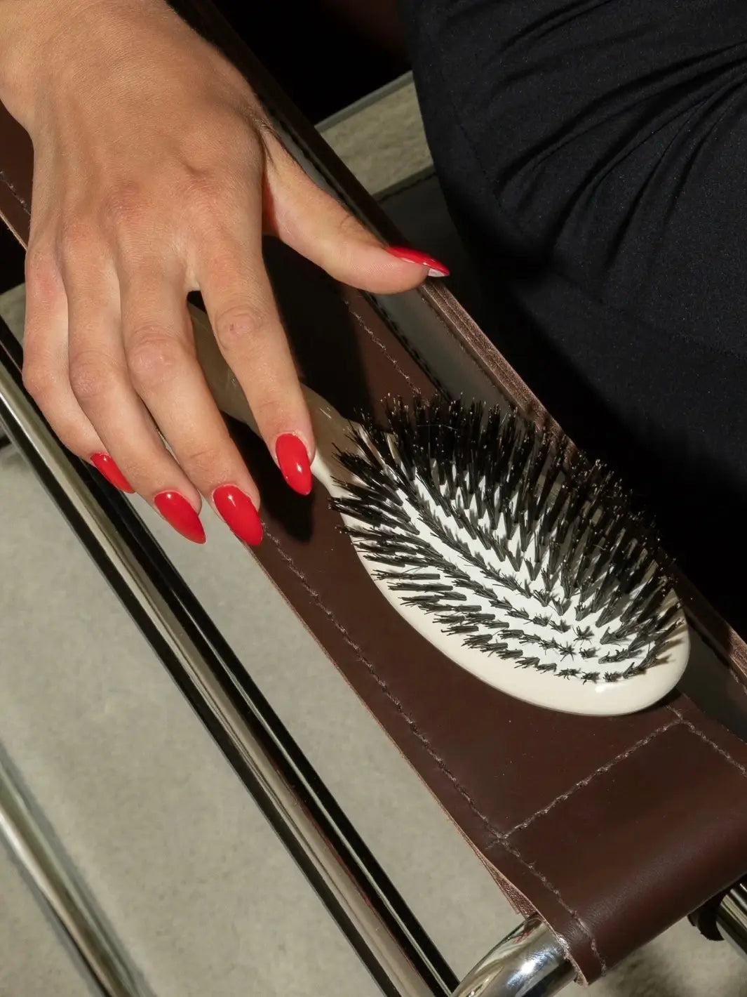 A hand with bright red almond-shaped nails rests on the La Bonne Brosse N.01 The Shine & Care Hairbrush The Universal Brush in Creamy White, displayed on a brown leather strap | Twentyseven Toronto