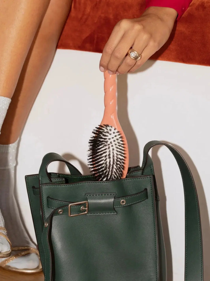 A lifestyle image showing the La Bonne Brosse N.01 The Shine & Care Hairbrush The Universal Brush in Coral Orange being placed into a dark green leather handbag | Twentyseven Toronto