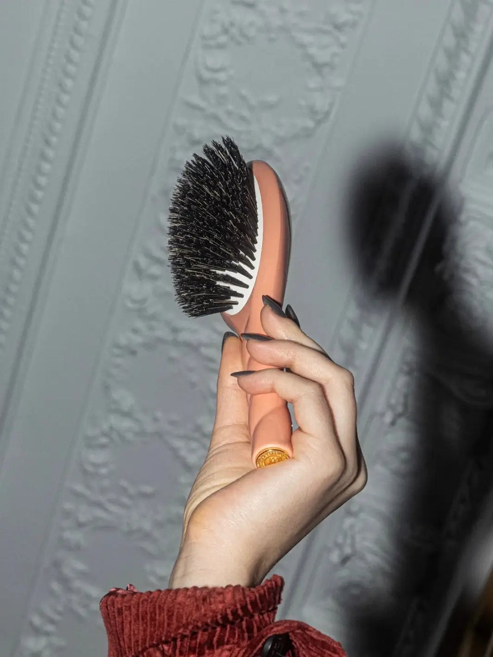 A hand holds the La Bonne Brosse N.01 The Shine & Care Hairbrush The Universal Brush in Coral Orange against a decorative white wall | Twentyseven Toronto