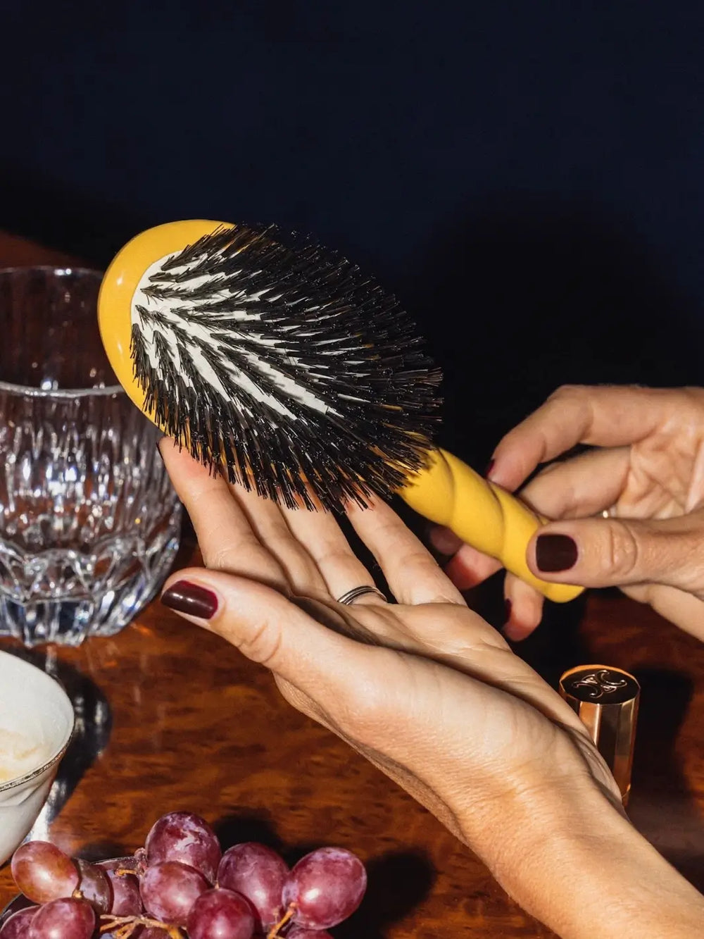 A person with deep burgundy nails holds the La Bonne Brosse N.01 The Shine & Care Hairbrush The Universal Brush in Saffron Yellow over a wooden table | Twentyseven Toronto
