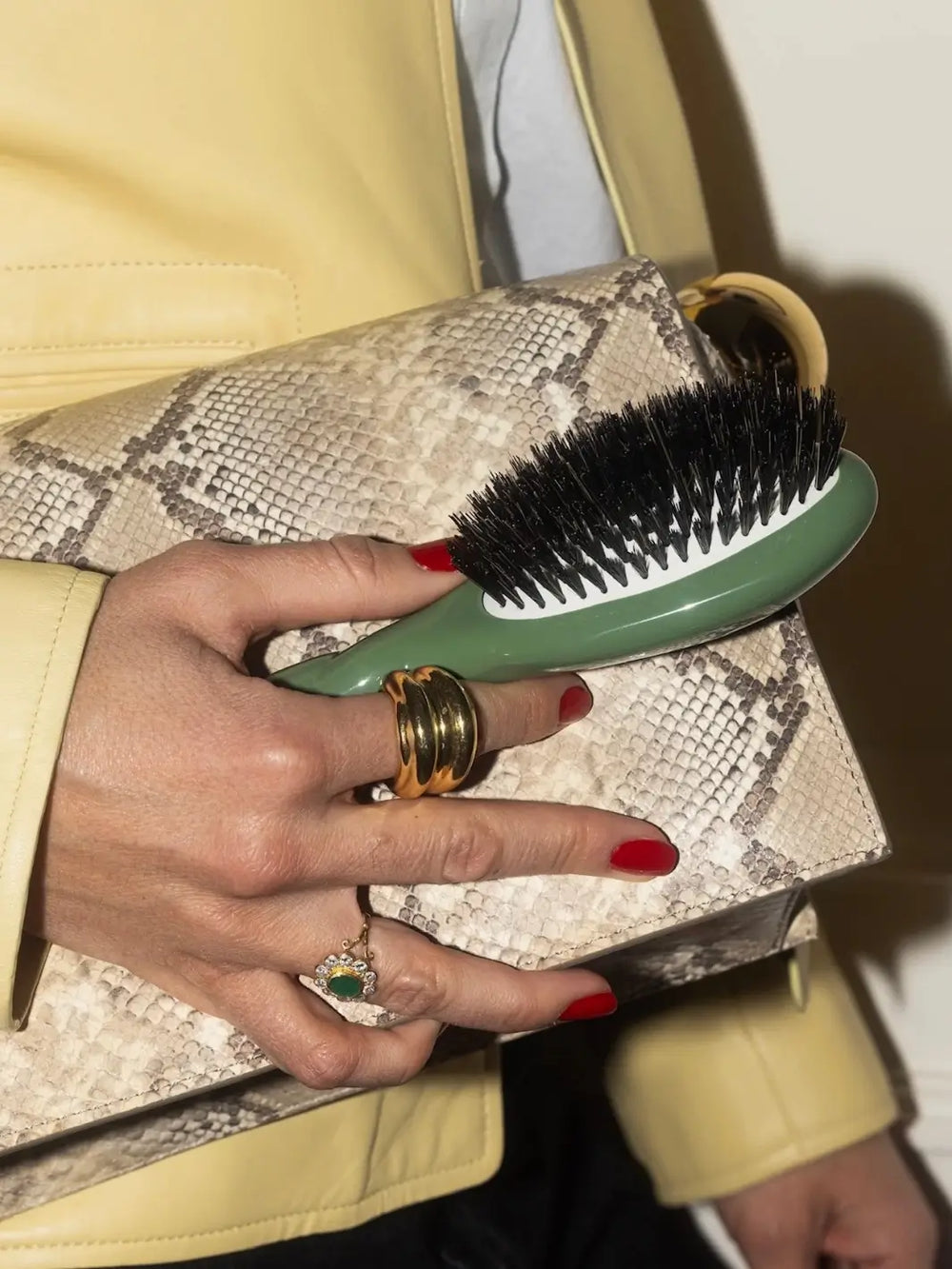 Hand with red nails and gold rings holds the La Bonne Brosse N.01 Small The Shine & Care Hairbrush The Universal Brush in Almond Green against a neutral snakeskin clutch and a pale yellow leather jacket | Twentyseven Toronto