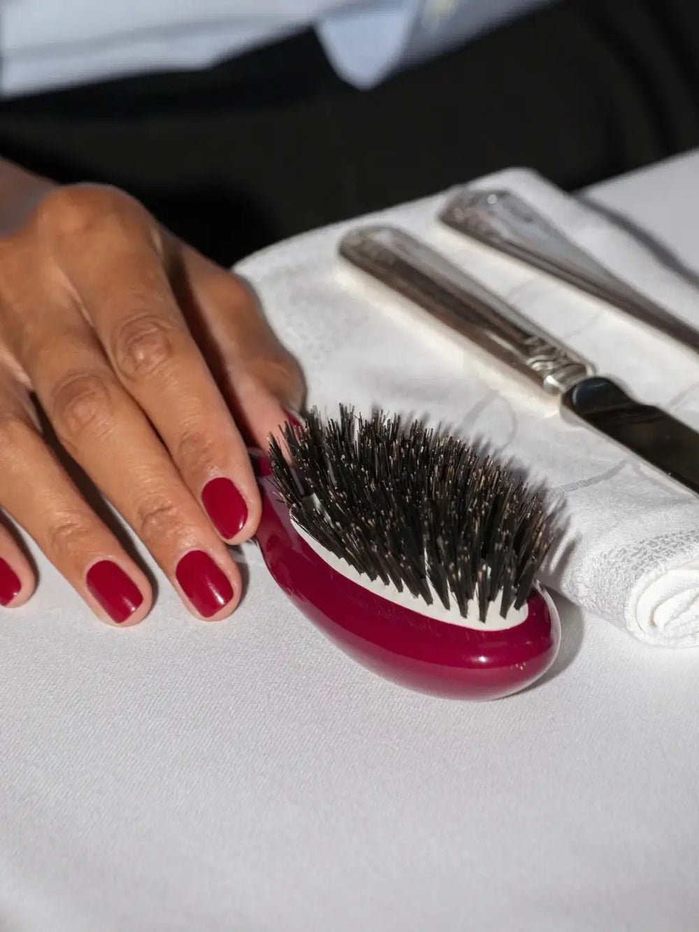 Hand with deep red manicured nails rests next to the La Bonne Brosse N.01 Small The Shine & Care Hairbrush The Universal Brush in Cherry Red on a white tablecloth | Twentyseven Toronto