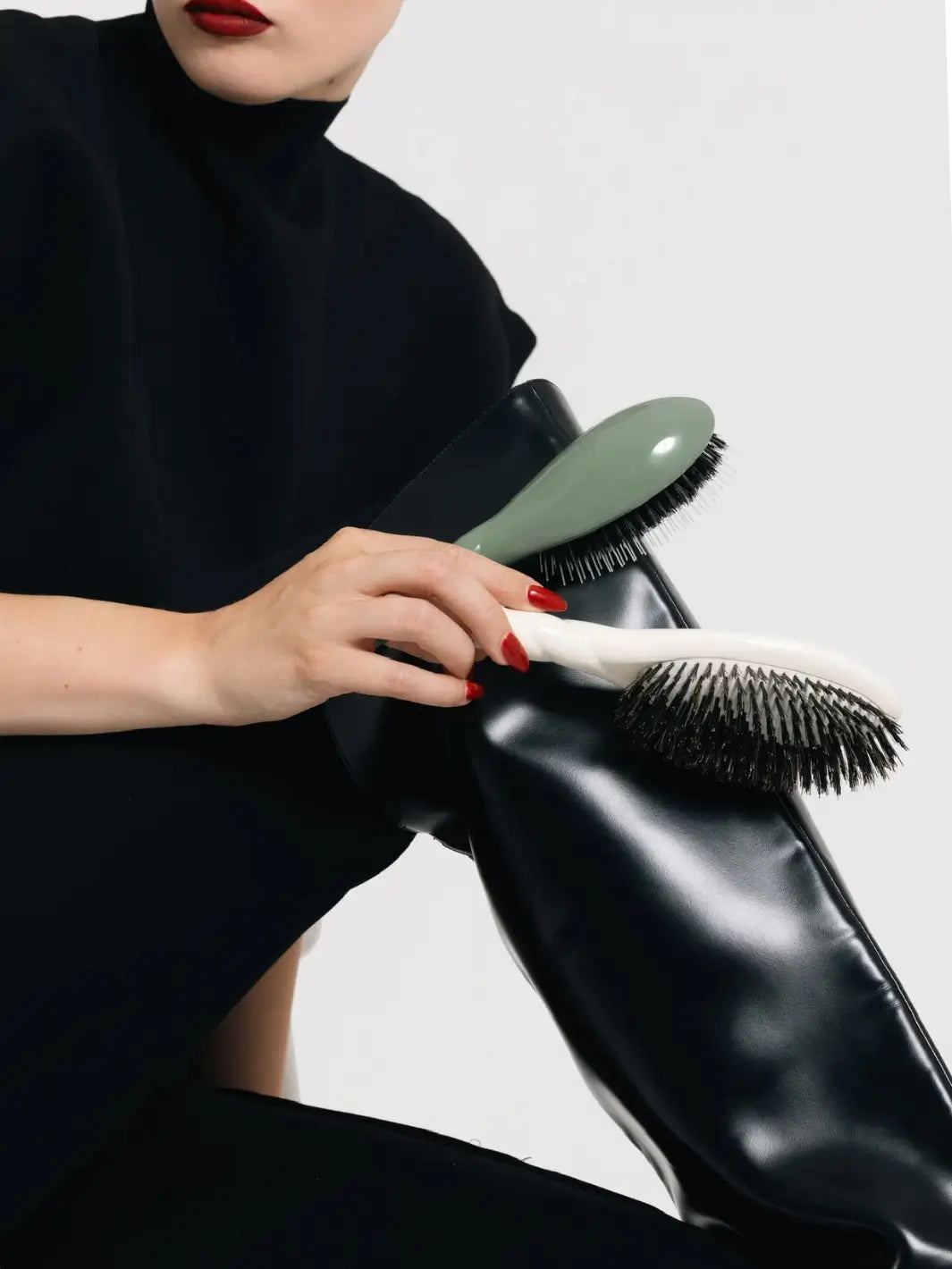 A person in a black high-neck top holds two hairbrushes against a black leather boot, featuring the Le Bonne Brosse N.02 Large The Essential Detangling & Conditioning Brush in Almond Green and Creamy White | Twentyseven Toronto