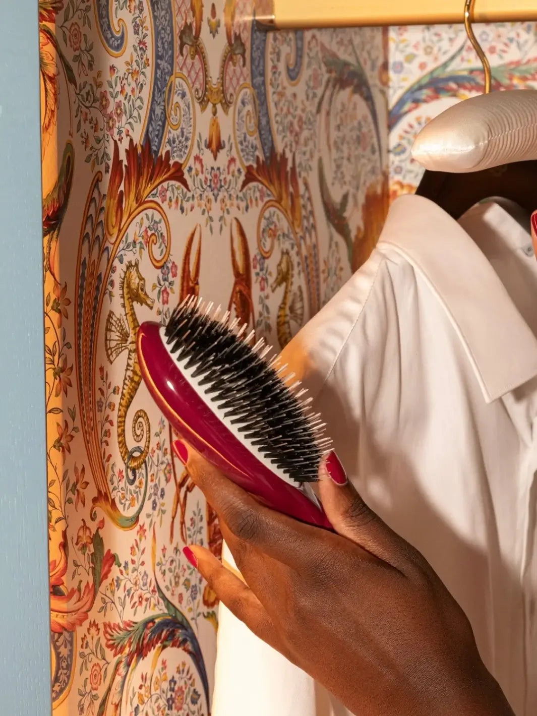 A person holds the N.02 Large The Le Bonne Brosse Essential Detangling & Conditioning Brush in Cherry Red in front of a decorative patterned wallpaper and a white collared shirt on a hanger | Twentyseven Toronto