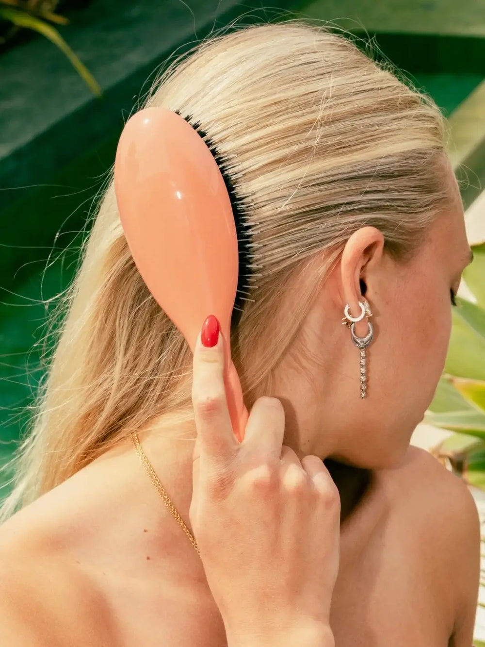 A side view of a blonde woman using the Le Bonne Brosse N.02 Large The Essential Detangling & Conditioning Brush in Orange Coral to smooth her hair | Twentyseven Toronto