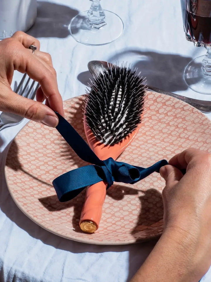 A lifestyle shot featuring the Le Bonne Brosse N.02 Large The Essential Detangling & Conditioning Brush in Orange Coral placed on a patterned dinner plate and being tied with a dark blue ribbon | Twentyseven Toronto