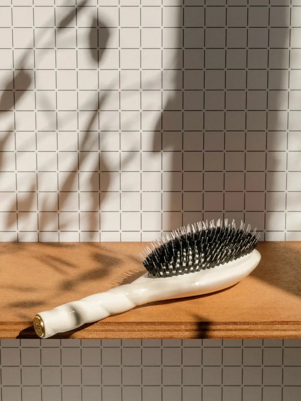 A shot of the Le Bonne Brosse N.02 Large The Essential Detangling & Conditioning Brush in Creamy White resting on a wooden shelf against a grid-patterned wall, with soft botanical shadows | Twentyseven Toronto