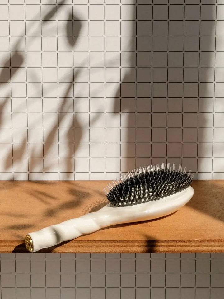 A shot of the Le Bonne Brosse N.02 Large The Essential Detangling & Conditioning Brush in Creamy White resting on a wooden shelf against a grid-patterned wall, with soft botanical shadows | Twentyseven Toronto