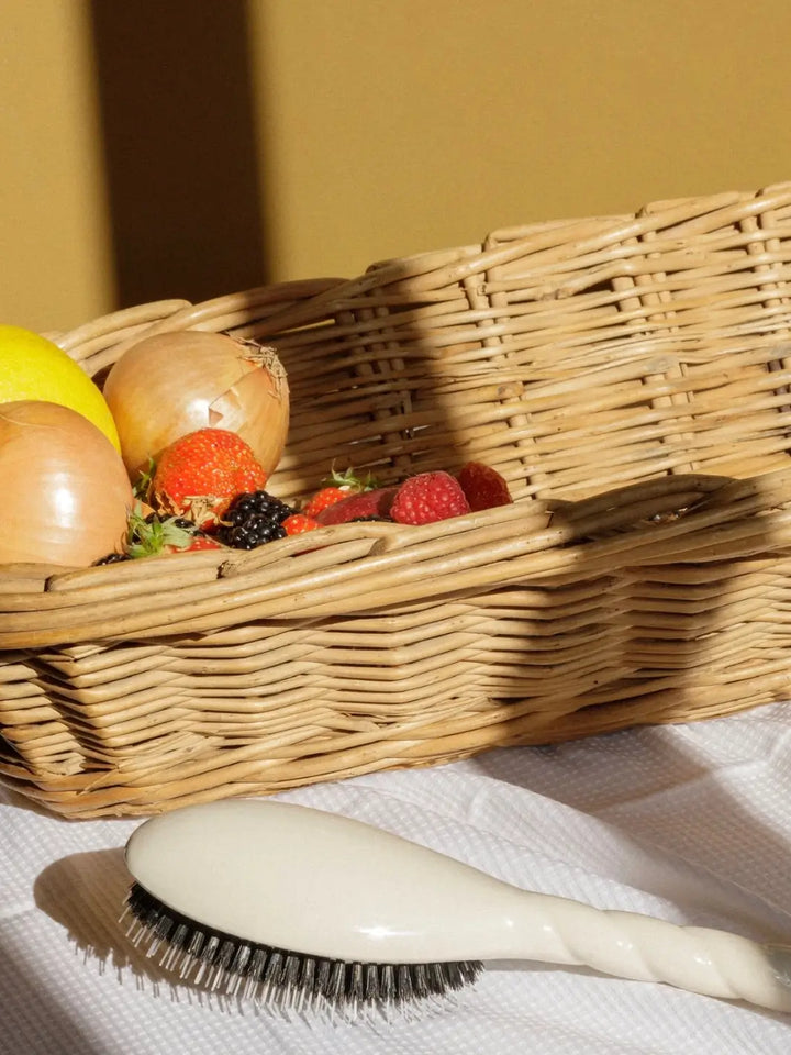 The Le Bonne Brosse N.02 Large The Essential Detangling & Conditioning Brush in Creamy White lying on a white textured cloth next to a wicker basket filled with fresh fruit and vegetables | Twentyseven Toronto