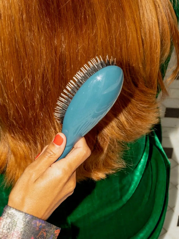 Person with red nails and long auburn hair using La Bonne Brosse N.02 Large The Essential Detangling & Conditioning Brush in Ink Blue against a green garment | Twentyseven Toronto