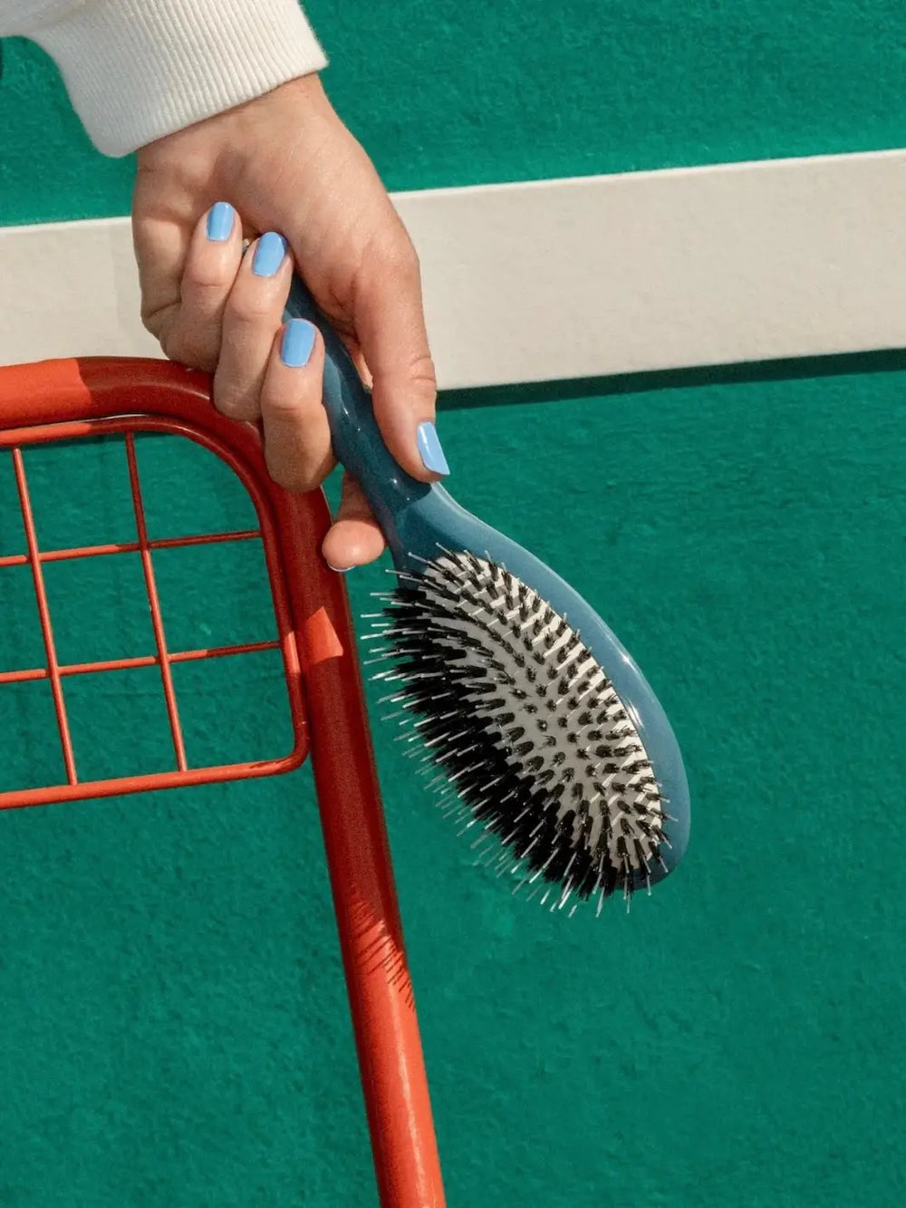 Hand with light blue nail polish holds the La Bonne Brosse N.02 Large The Essential Detangling & Conditioning Brush in Ink Blue against a vibrant teal wall and a red metal chair | Twentyseven Toronto