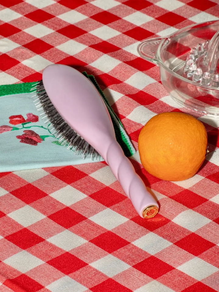 The La Bonne Brosse N.03 Small The Essential Softness & Sensitive Scalp Brush in Lilac Pink rests on a red and white checkered tablecloth next to an orange and a glass juicer | Twentyseven Toronto
