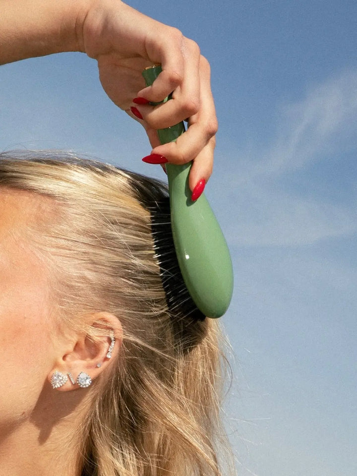 A close-up shot of a blonde woman using the La Bonne Brosse N.02 Small The Essential Detangling & Conditioning Brush in Almond Green against a clear blue sky | Twentyseven Toronto