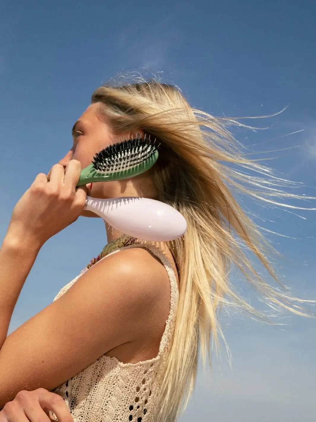 An outdoor photo of a blonde woman with wind-swept hair holding the La Bonne Brosse N.02 Small The Essential Detangling & Conditioning Brush in Almond Green and Lilac Pink against a bright blue sky | Twentyseven Toronto