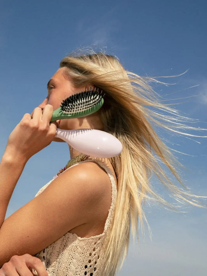 An outdoor photo of a blonde woman with wind-swept hair holding the La Bonne Brosse N.02 Small The Essential Detangling & Conditioning Brush in Almond Green and Lilac Pink against a bright blue sky | Twentyseven Toronto