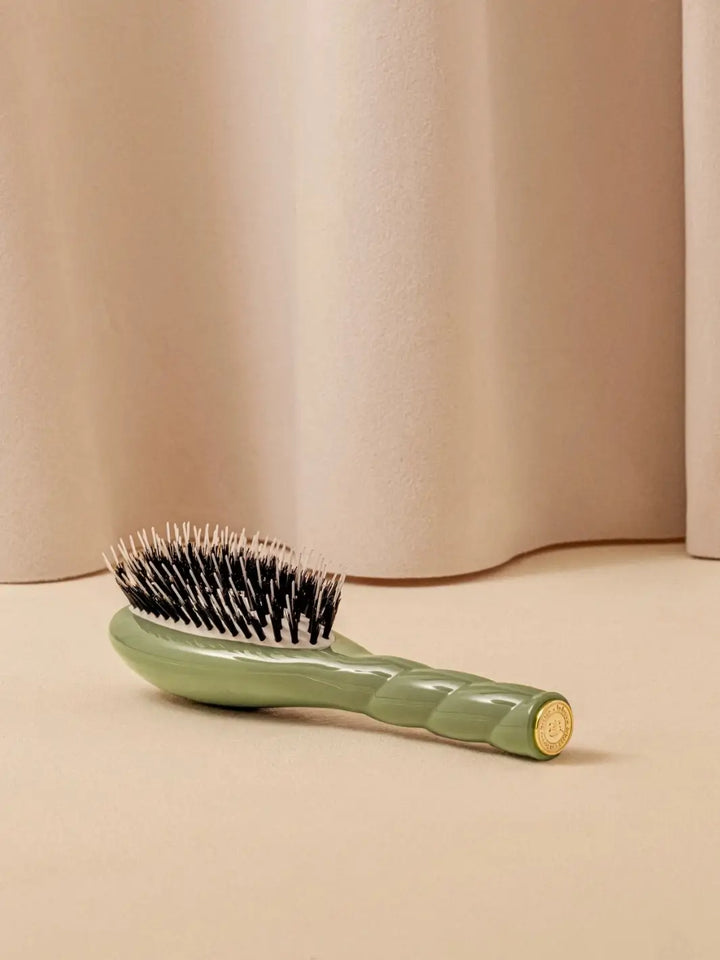 A shot of the La Bonne Brosse N.02 Small The Essential Detangling & Conditioning Brush in Almond Green resting on a neutral surface against a soft draped curtain background | Twentyseven Toronto