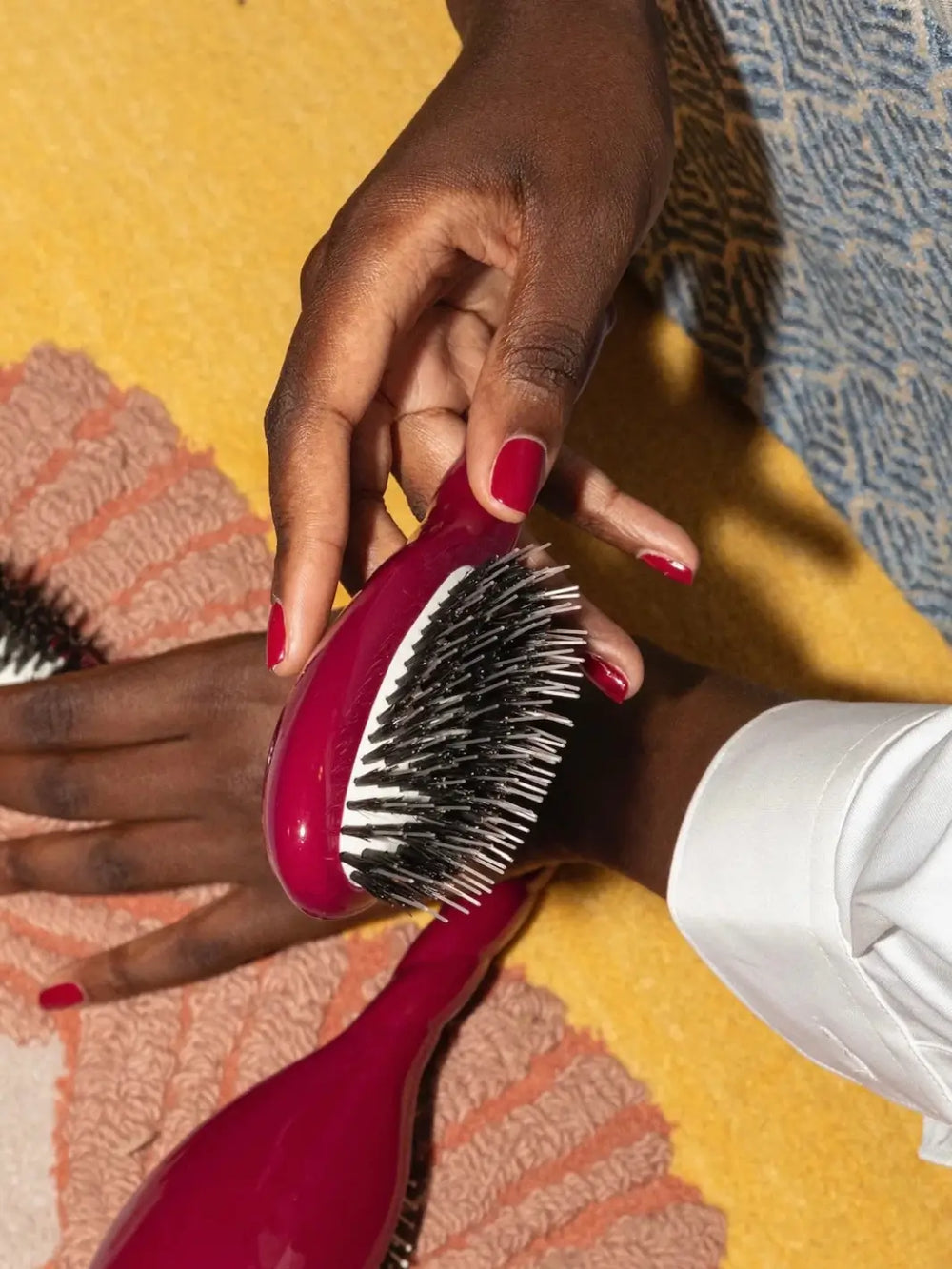 A hand with red nails holding the La Bonne Brosse N.02 Small The Essential Detangling & Conditioning Brush in Cherry Red over a yellow and pink textured surface | Twentyseven Toronto