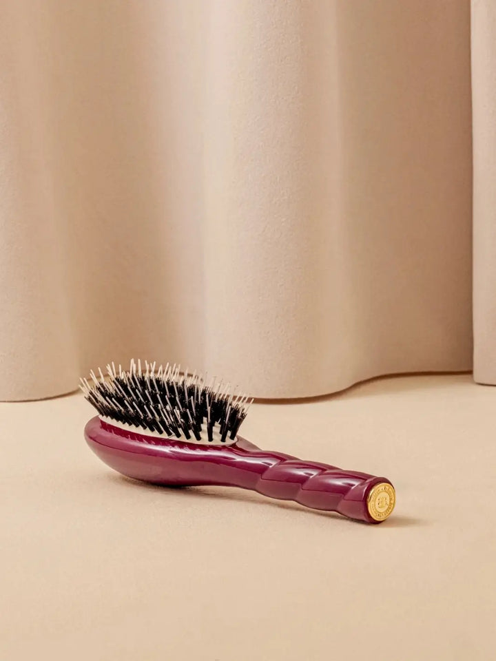 A shot of the La Bonne Brosse N.02 Small The Essential Detangling & Conditioning Brush in Cherry Red resting on a neutral surface against a soft draped curtain background | Twentyseven Toronto