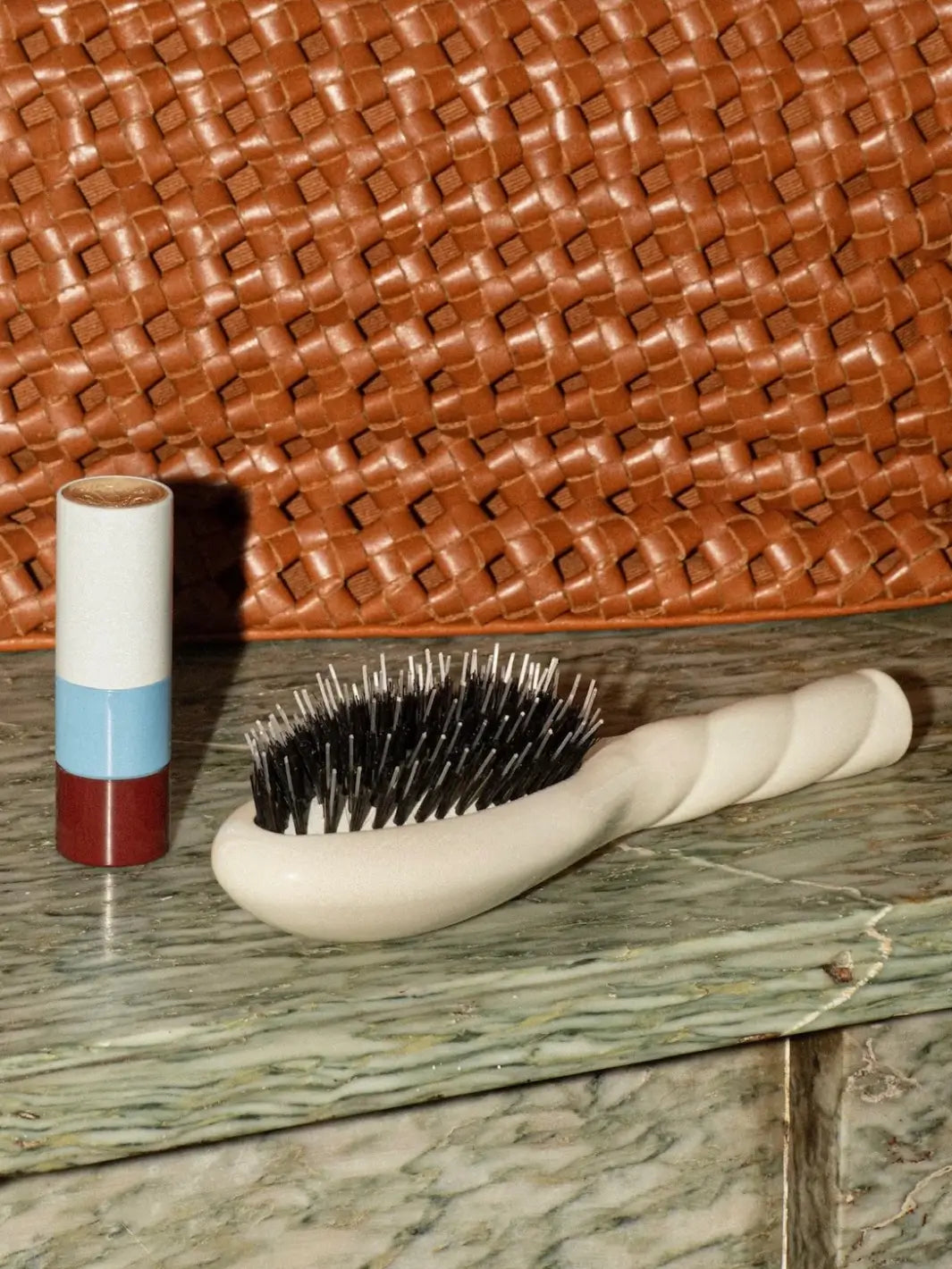 La Bonne Brosse N.02 Small The Essential Detangling & Conditioning Hairbrush in Creamy White resting on a green surface next to a tricolor lipstick, with a woven brown leather bag in the background | Twentyseven Toronto