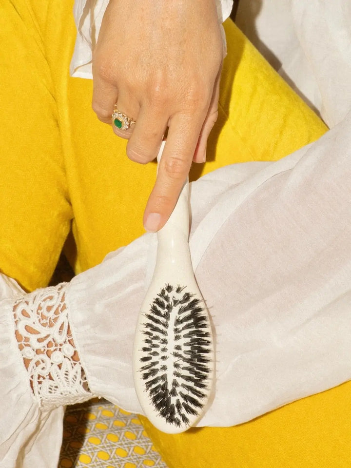 Person wearing a white lace-cuffed blouse and yellow trousers holding the La Bonne Brosse N.02 Small The Essential Detangling & Conditioning Brush in Creamy White | Twentyseven Toronto