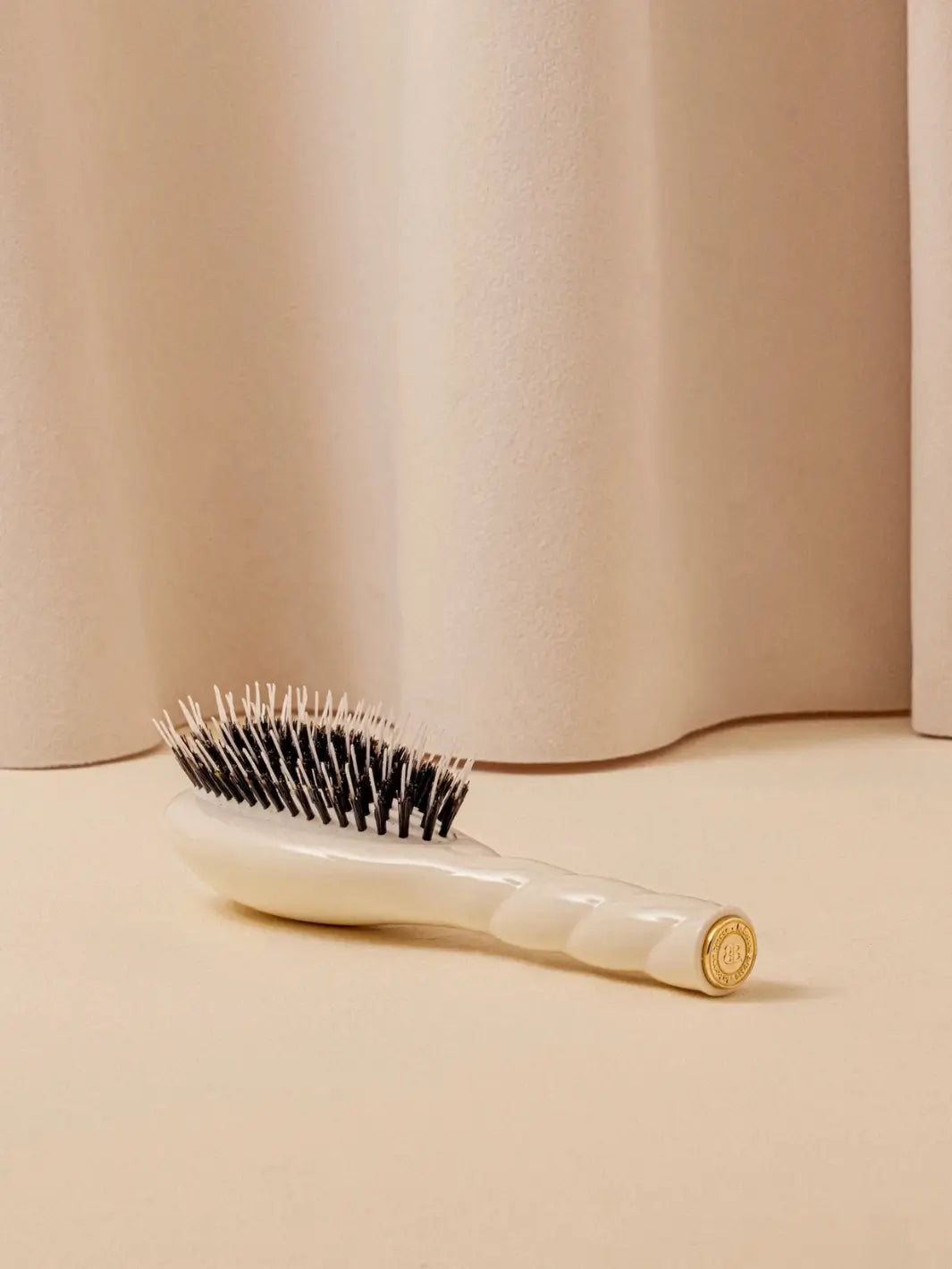 A shot of the La Bonne Brosse N.02 Small The Essential Detangling & Conditioning Brush in Creamy White resting on a neutral surface against a soft draped curtain background | Twentyseven Toronto