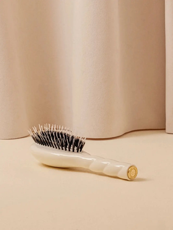 A shot of the La Bonne Brosse N.02 Small The Essential Detangling & Conditioning Brush in Creamy White resting on a neutral surface against a soft draped curtain background | Twentyseven Toronto