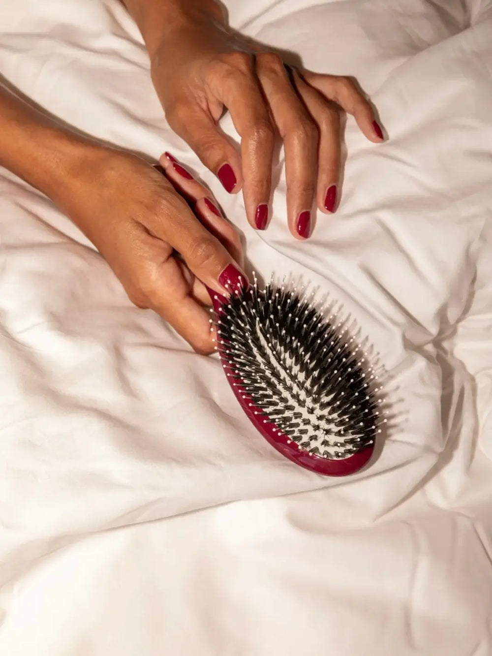 A pair of hands with cherry red manicured nails holds the La Bonne Brosse N.03 Large The Essential Softness & Sensitive Scalp Brush in Cherry Red against white bedding | Twentyseven Toronto