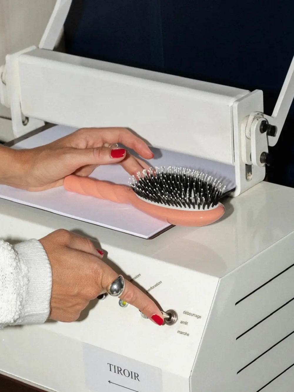La Bonne Brosse N.03 Large The Essential Softness & Sensitive Scalp Brush in Orange Coral placed on a white industrial machine while a person with a silver ring and red nail polish operates a toggle switch | Twentyseven Toronto