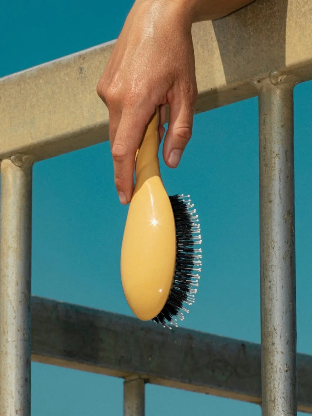 Hand holding the La Bonne Brosse N.03 Large The Essential Softness & Sensitive Scalp Brush in Saffron Yellow over a metal railing against a clear blue sky | Twentyseven Toronto