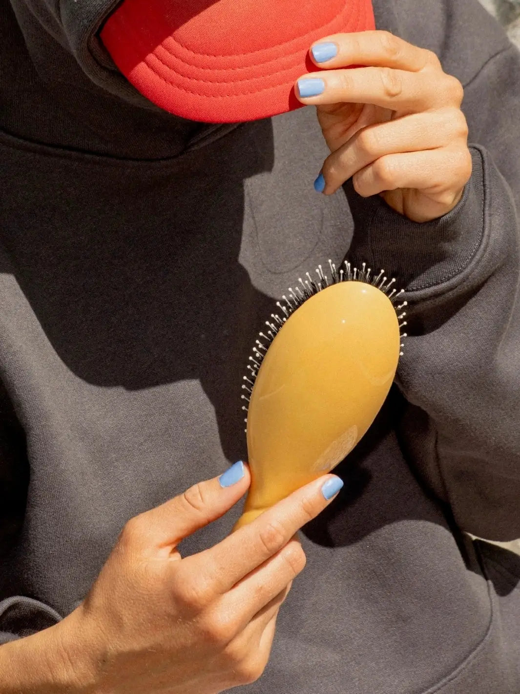 Person with light blue nail polish and a red cap holds the La Bonne Brosse N.03 Large The Essential Softness & Sensitive Scalp Brush in Saffron Yellow against a dark grey hoodie | Twentyseven Toronto