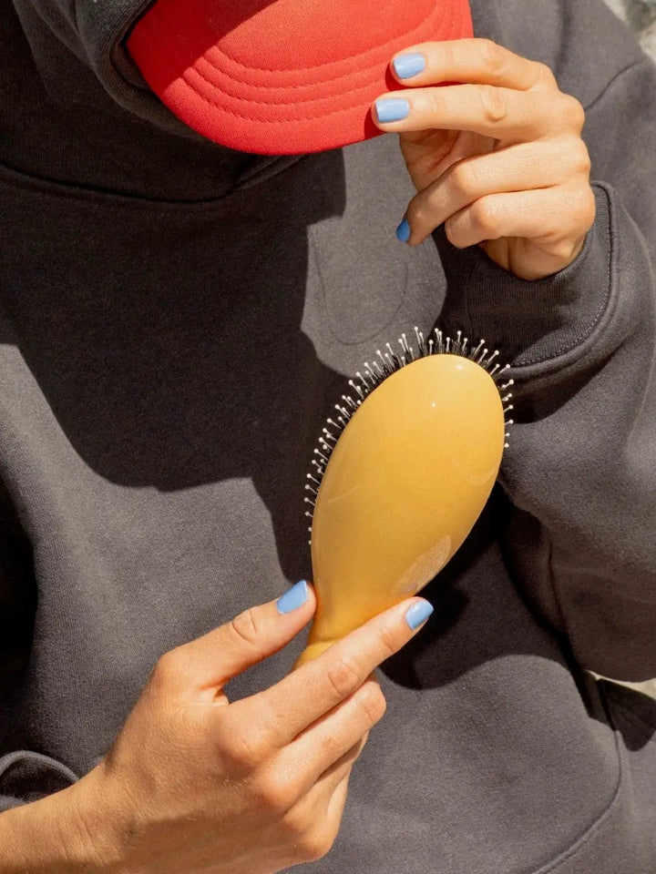 Person with light blue nail polish and a red cap holds the La Bonne Brosse N.03 Large The Essential Softness & Sensitive Scalp Brush in Saffron Yellow against a dark grey hoodie | Twentyseven Toronto