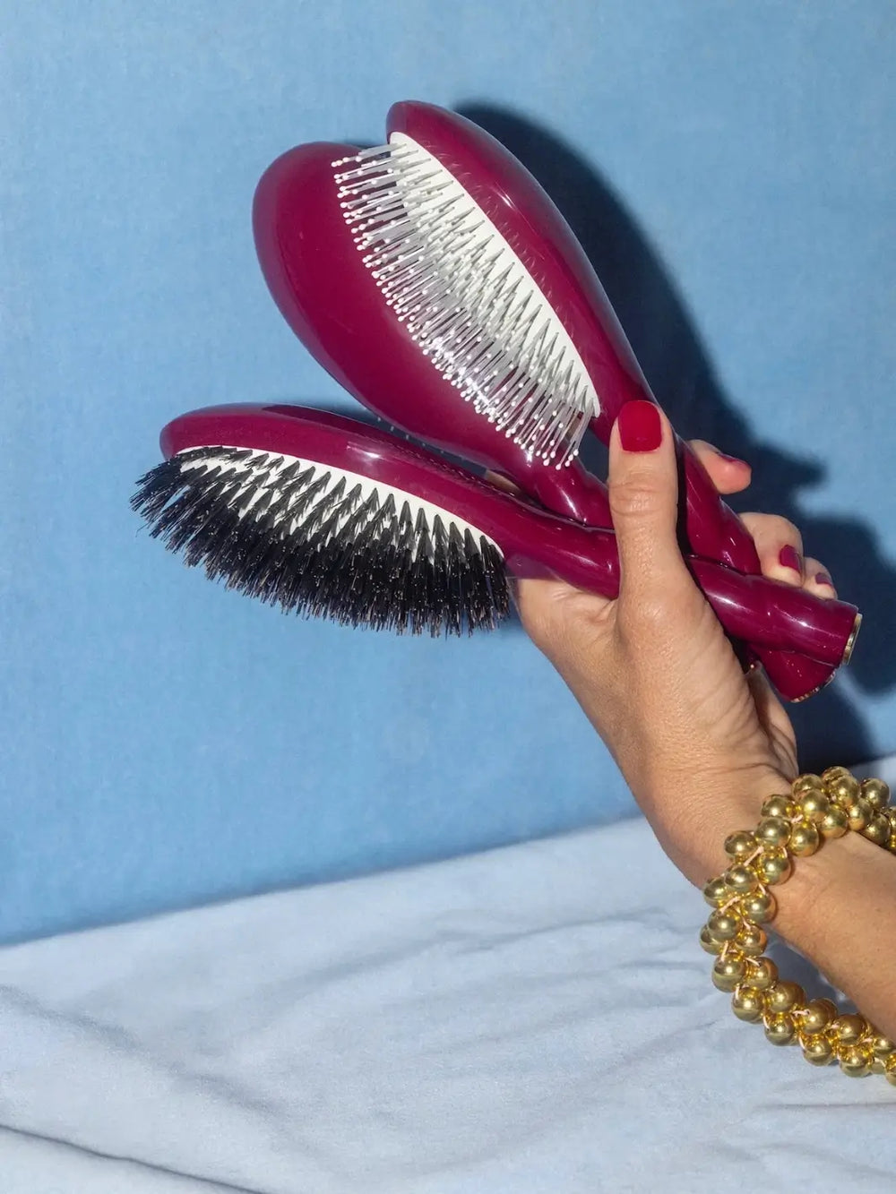 Hand with red nails and a gold beaded bracelet holds three La Bonne Brosse brushes including N.04 Large The Detangling & Massaging Miracle Brush in Cherry Red against a blue background | Twentyseven Toronto