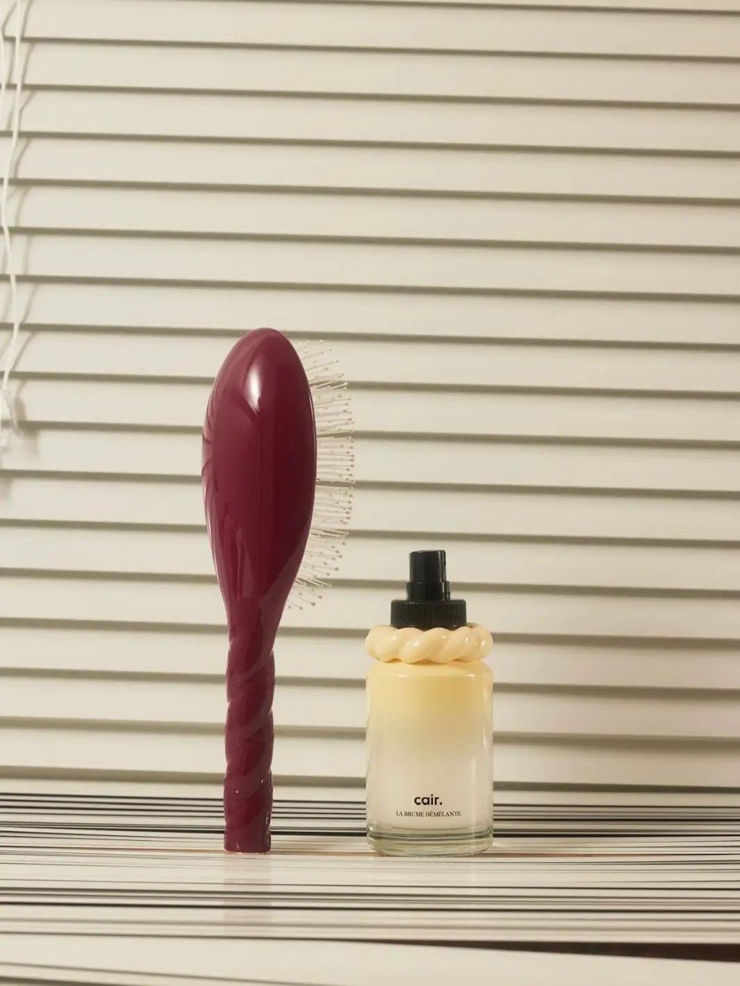 La Bonne Brosse N.04 Large The Detangling & Massaging Miracle Brush in Cherry Red stands vertically next to a bottle of Cair hair mist against a backdrop of white window blinds | Twentyseven Toronto