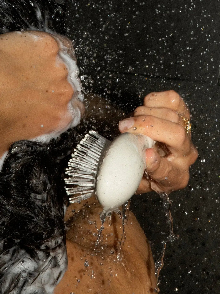 Under a stream of water, a hand holds the N.04 Small The Detangling & Massaging Miracle Brush in Creamy White while brushing through soapy, lathered hair | Twentyseven Toronto