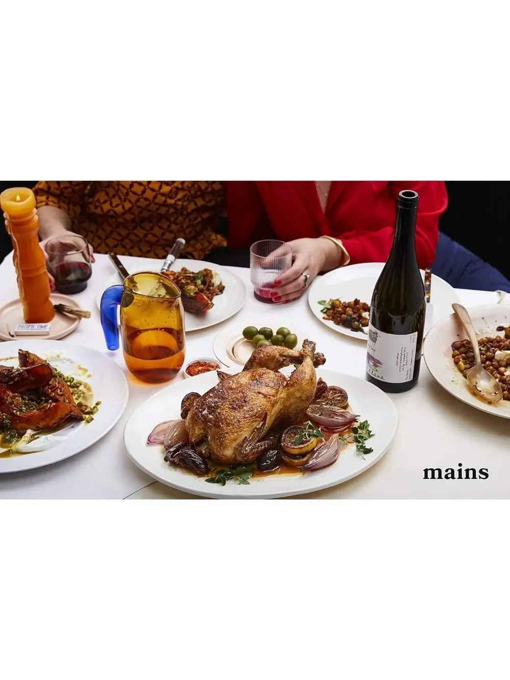 A dinner table spread features a platter with a whole roasted chicken surrounded by shallots and lemons, showcasing dishes from Nothing Fancy: Unfussy Food for Having People Over by Alison Roman | Twentyseven Toronto