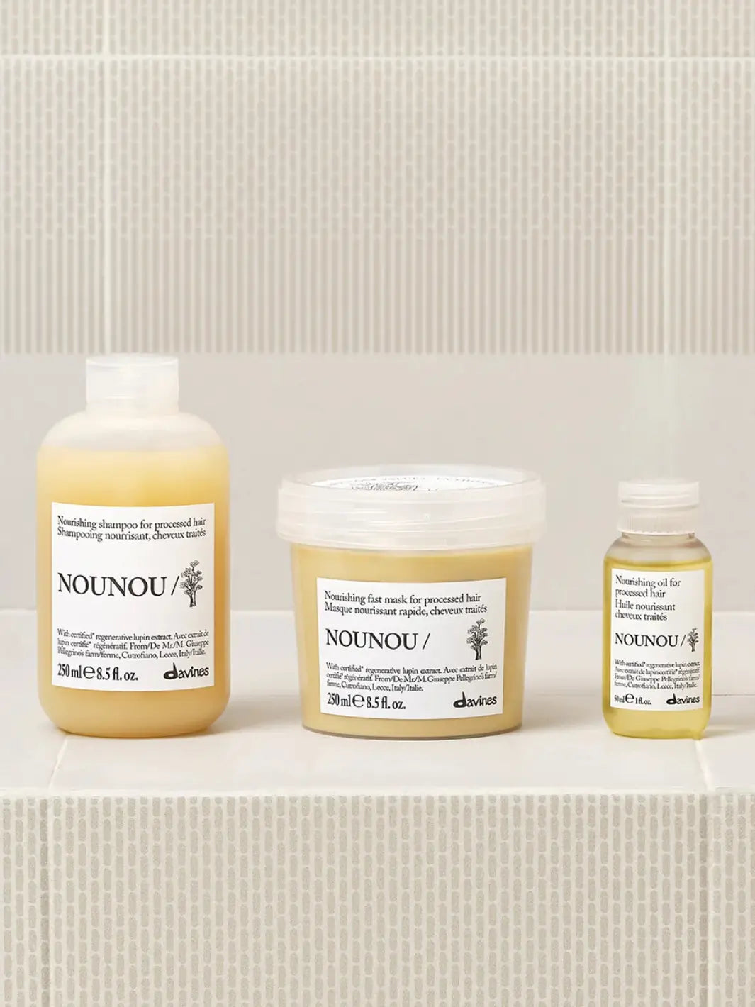 A product group shot shows the small bottle of Davines NOUNOU Oil standing on a light tiled ledge alongside its matching nourishing shampoo bottle and fast mask tub for processed hair | Twentyseven Toronto
