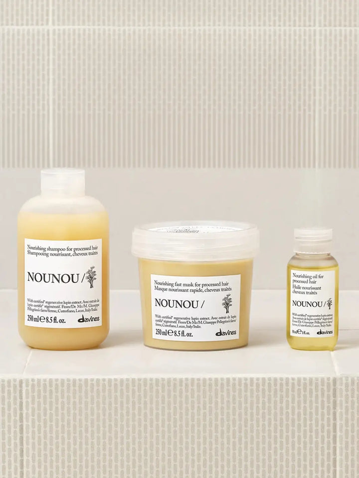 A product group shot shows the small bottle of Davines NOUNOU Oil standing on a light tiled ledge alongside its matching nourishing shampoo bottle and fast mask tub for processed hair | Twentyseven Toronto