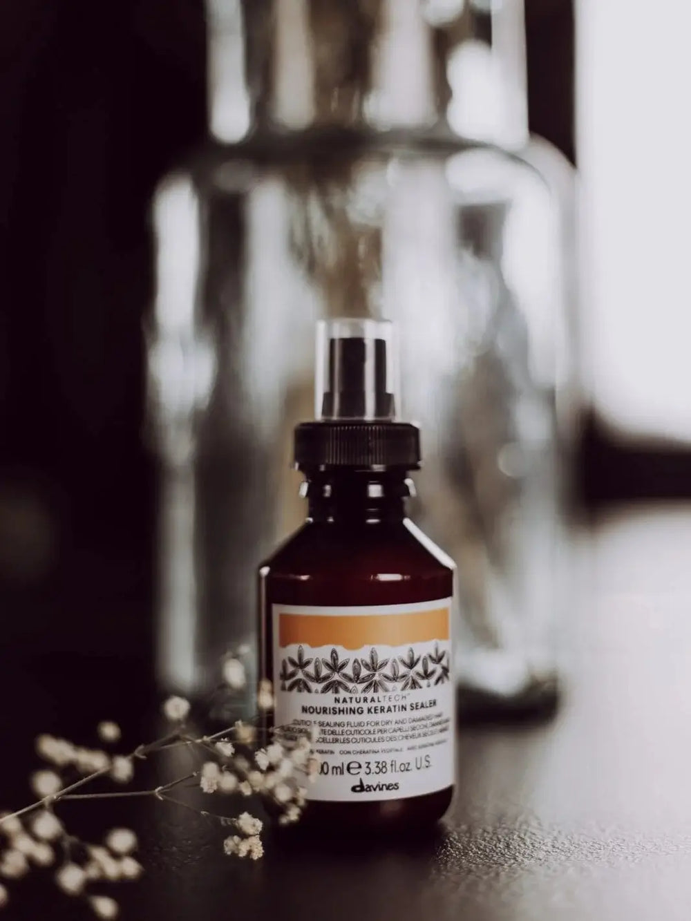 A spray bottle of Davines NOURISHING Keratin Sealer sits on a dark surface in front of a blurred glass jar, with a sprig of small white flowers | Twentyseven Toronto