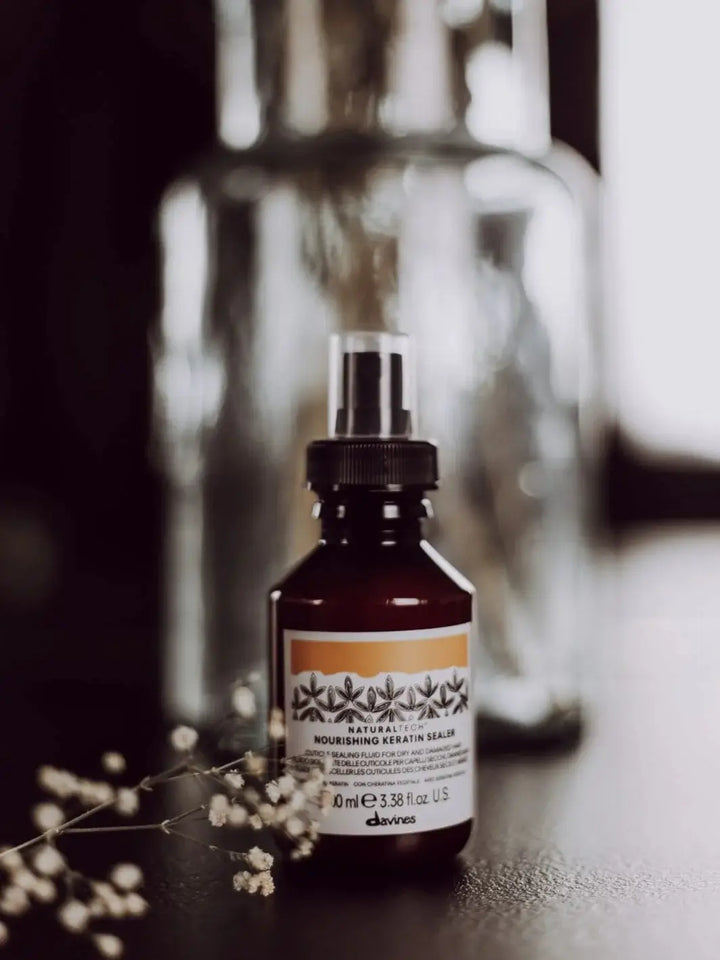 A spray bottle of Davines NOURISHING Keratin Sealer sits on a dark surface in front of a blurred glass jar, with a sprig of small white flowers | Twentyseven Toronto