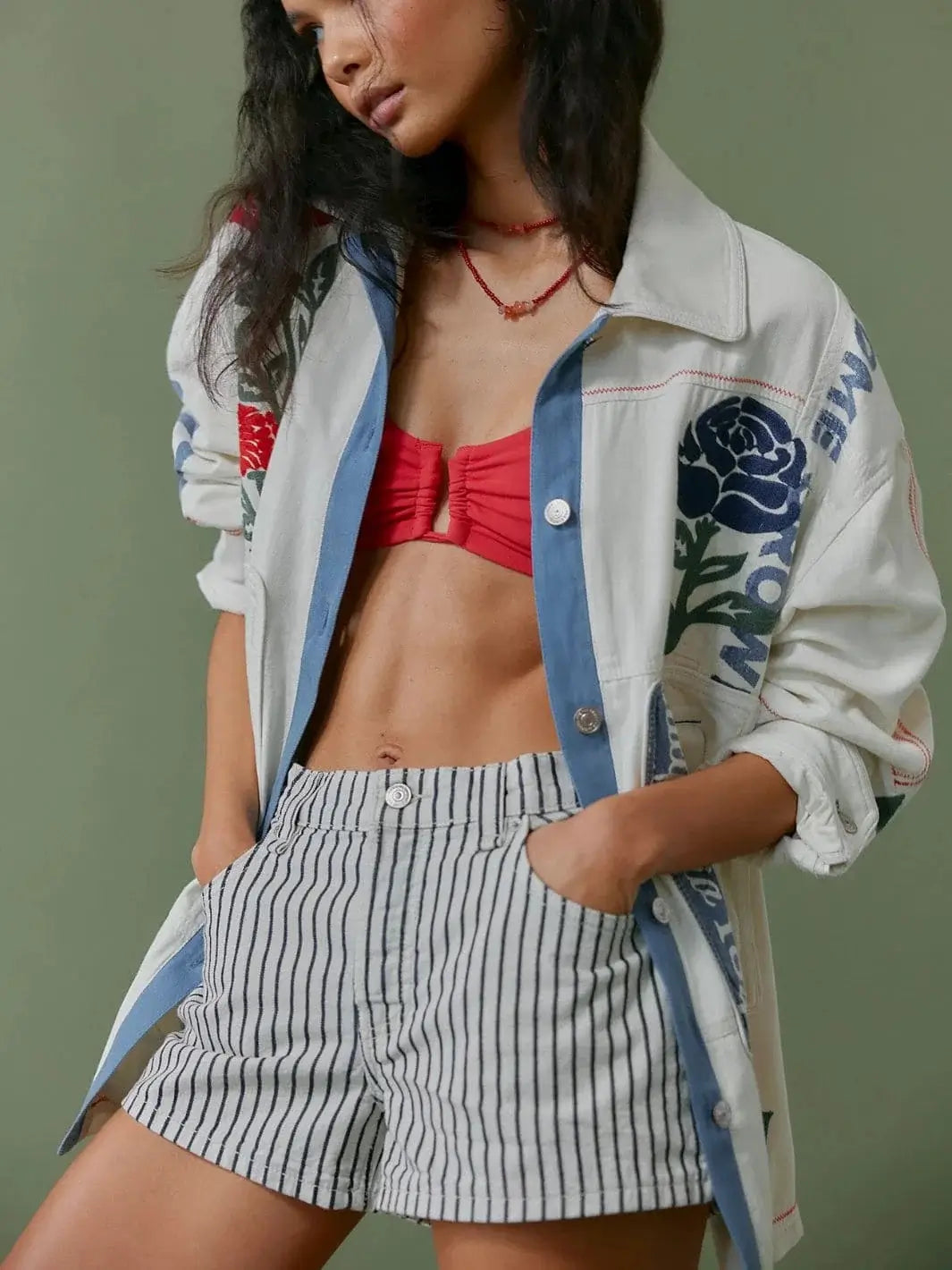 A model stands with her hands in her pockets showcasing the vertical stripes of the Free People Now Or Never Railroad Denim Shorts - Washed Railroad, styled with a red bralette and a graphic cream jacket | Twentyseven Toronto