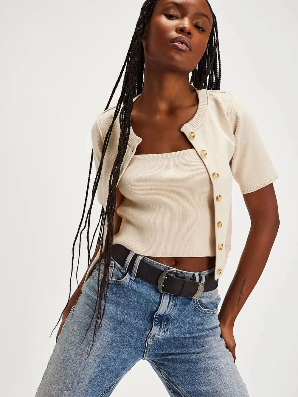 A front view shows a model with long braids wearing the Free People We The Free NY Twosie - Ecru styled as an unbuttoned, cropped knit set with light-wash jeans and a dark leather belt | Twentyseven Toronto