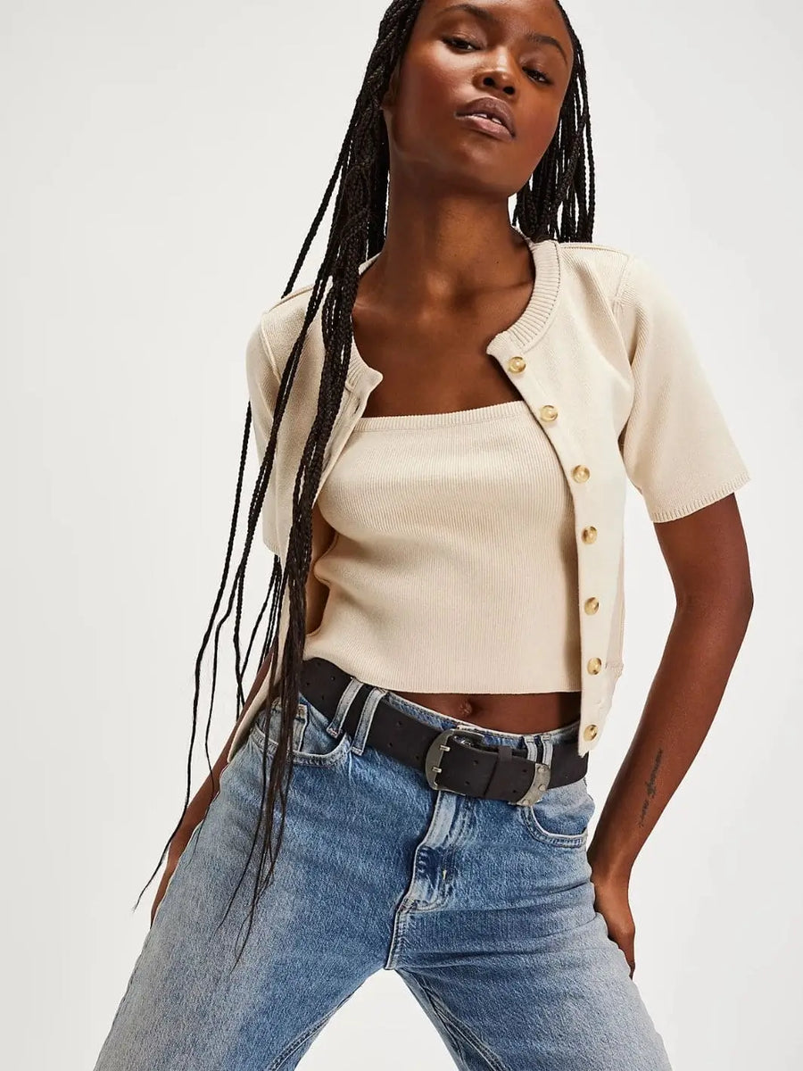 A front view shows a model with long braids wearing the Free People We The Free NY Twosie - Ecru styled as an unbuttoned, cropped knit set with light-wash jeans and a dark leather belt | Twentyseven Toronto