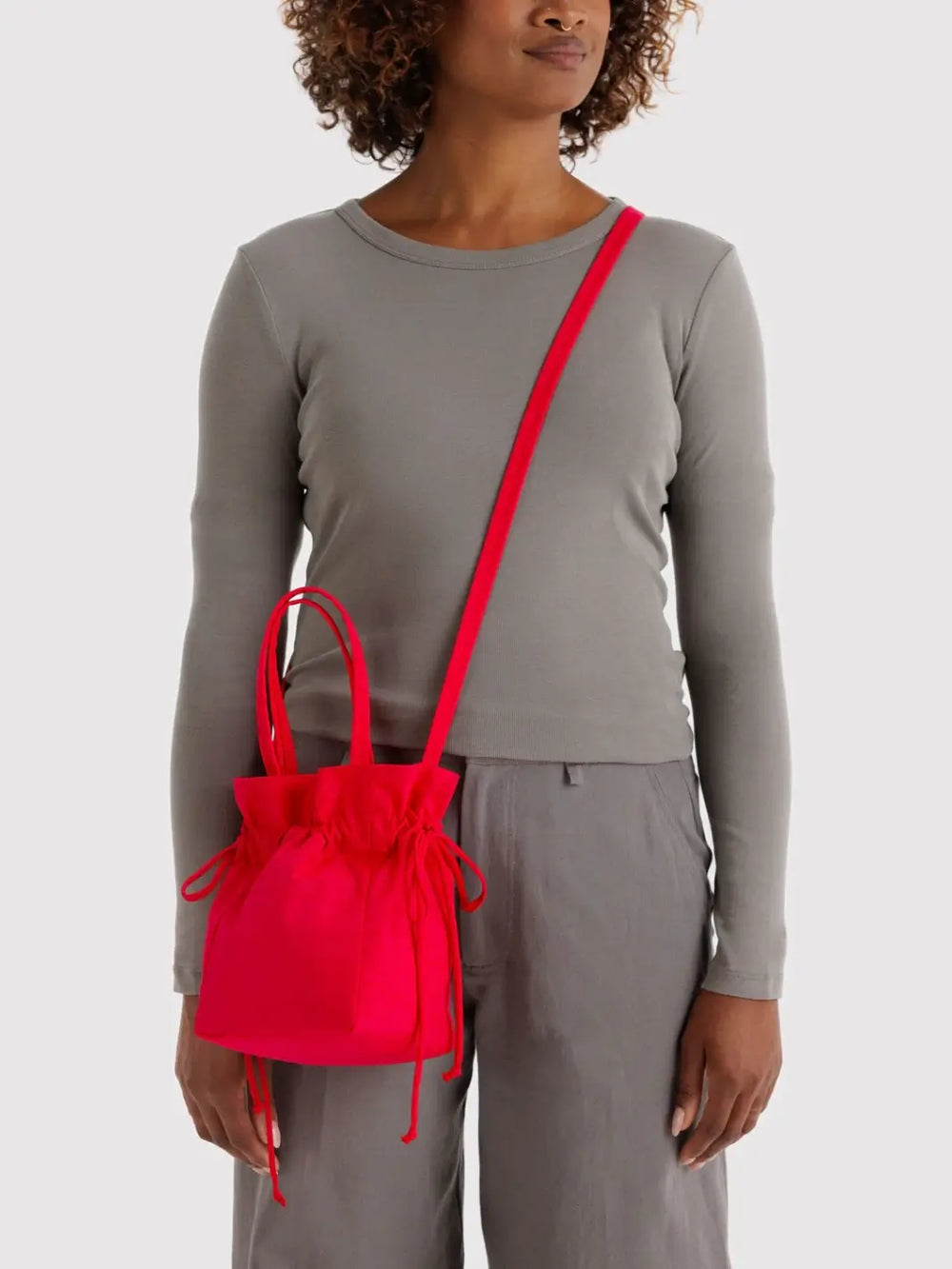 Person wears a BAGGU Nylon Drawstring Bag in Candy Apple as a crossbody over a gray long-sleeve shirt and matching trousers | Twentyseven Toronto