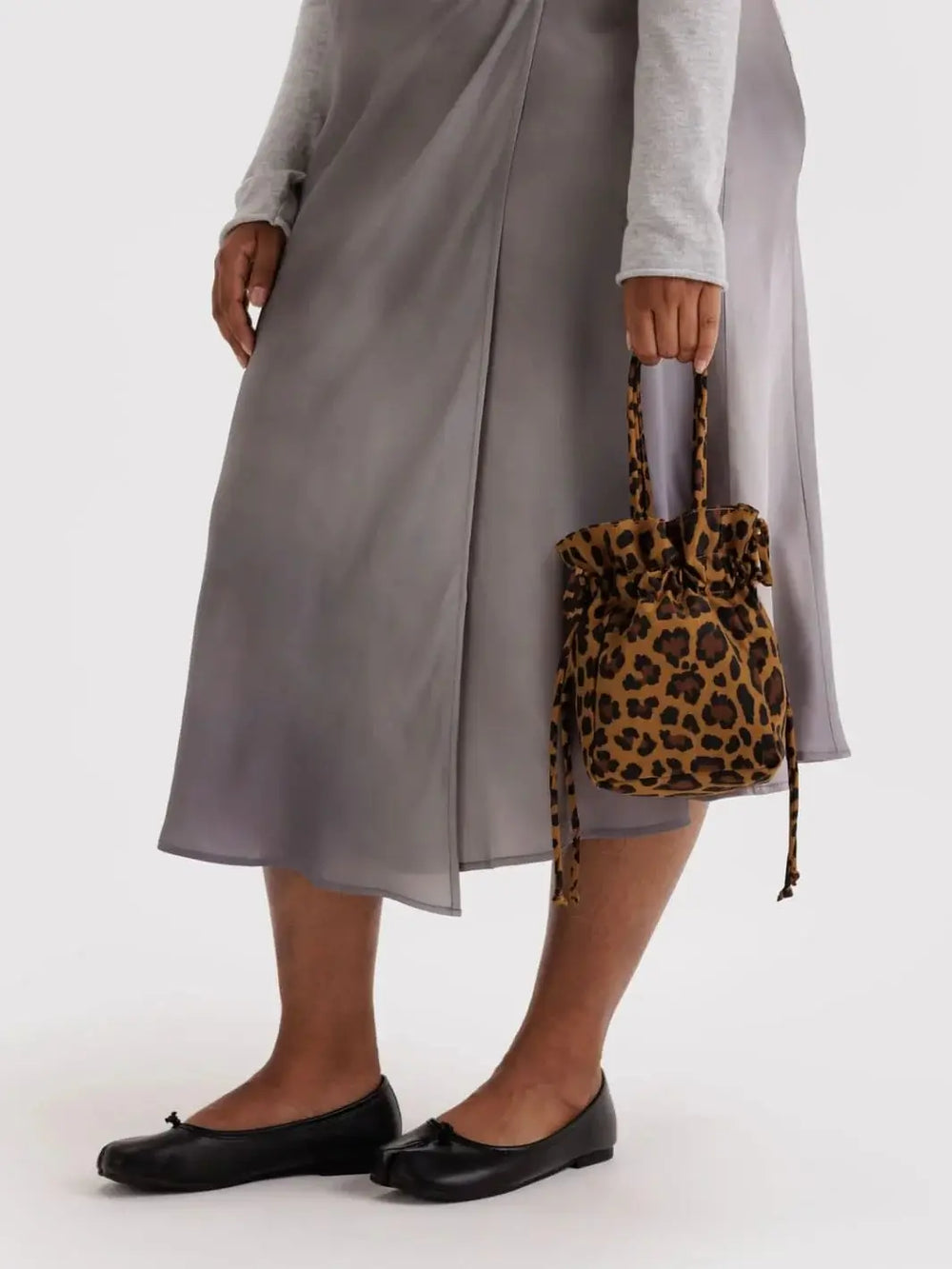 A mid-section view of a person wearing a grey satin skirt and black ballet flats, holding a Baggu Nylon Drawstring Bag in Leopard by its top handles | Twentyseven Toronto